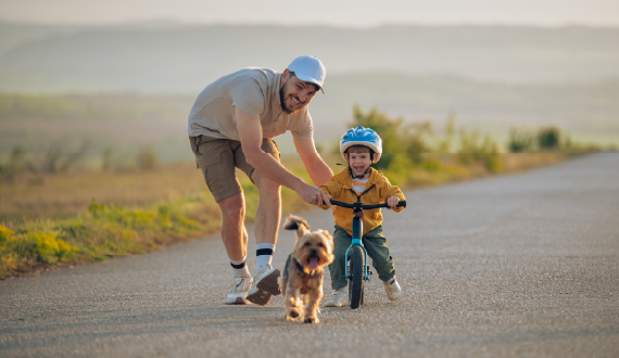 Smiling dad helping his toddler son ride a bike on a rural road at sunset with a small dog.