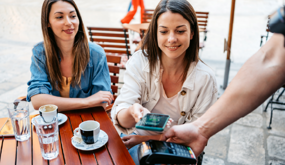 Two women sitting at a cafe and one tapping her phone to make a contactless payment.